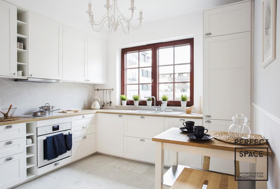 Jasna kuchnia z białymi meblami, drewnianym blatem i oknem w ciemnej ramie, na parapecie doniczki z ziołami, na stole czarne filiżanki i słoik.