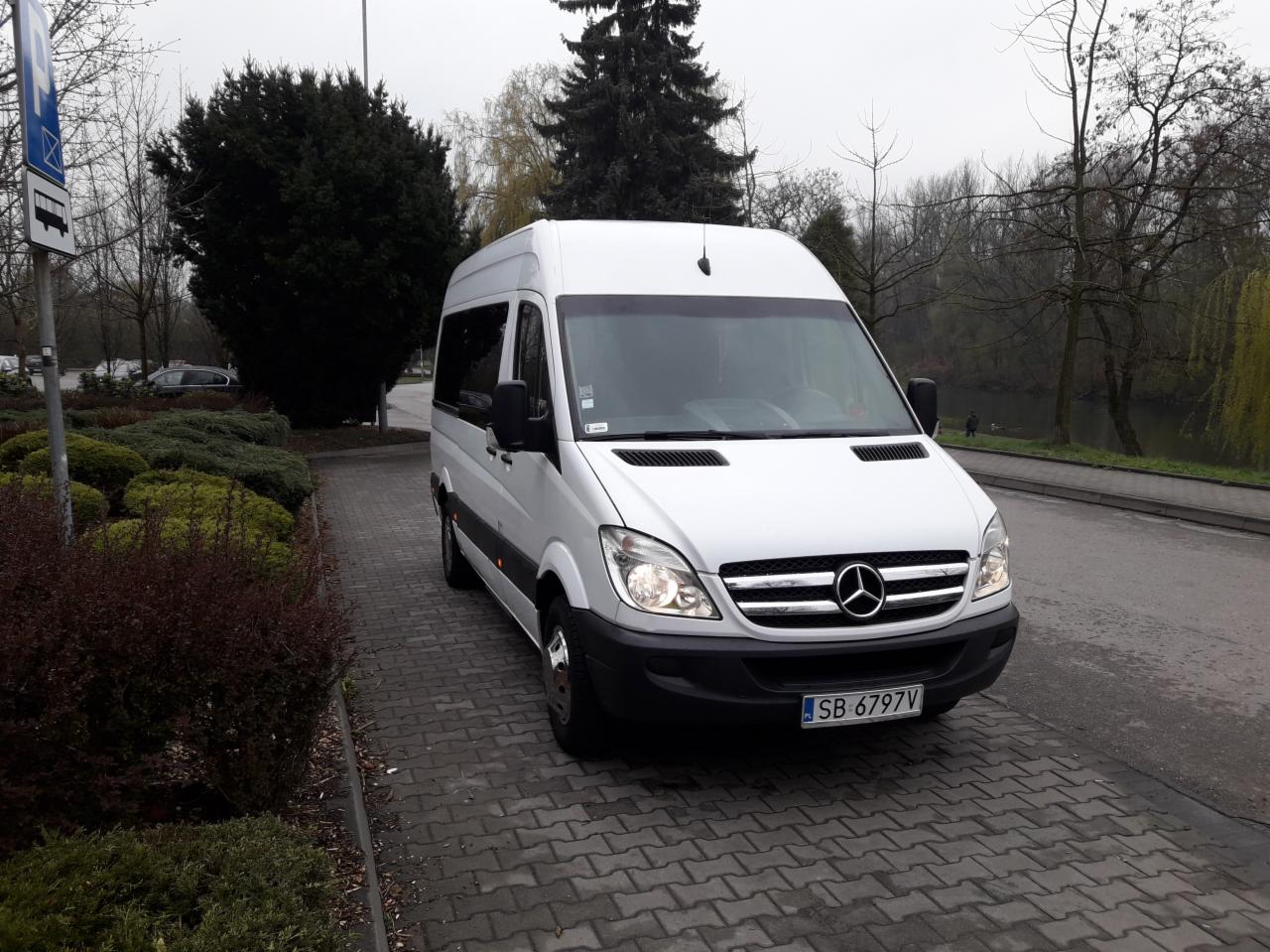 Biały bus Mercedes-Benz Sprinter z czarnymi szybami na parkingu z kostki brukowej, widoczna tablica rejestracyjna SB 6797V i znak przystanku w tle.