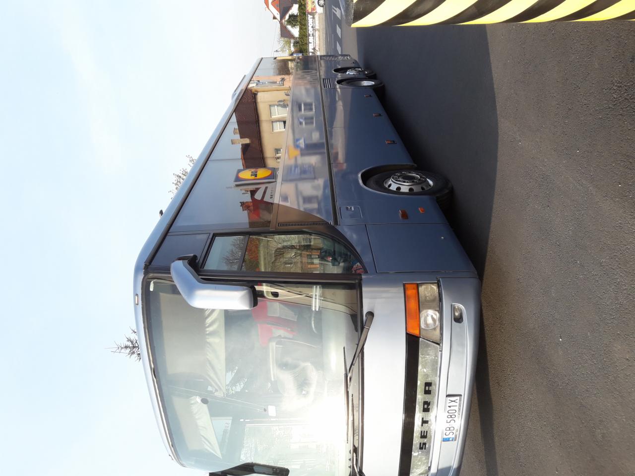 Szary autokar SETRA na parkingu z widocznym odbiciem budynku w szybach.