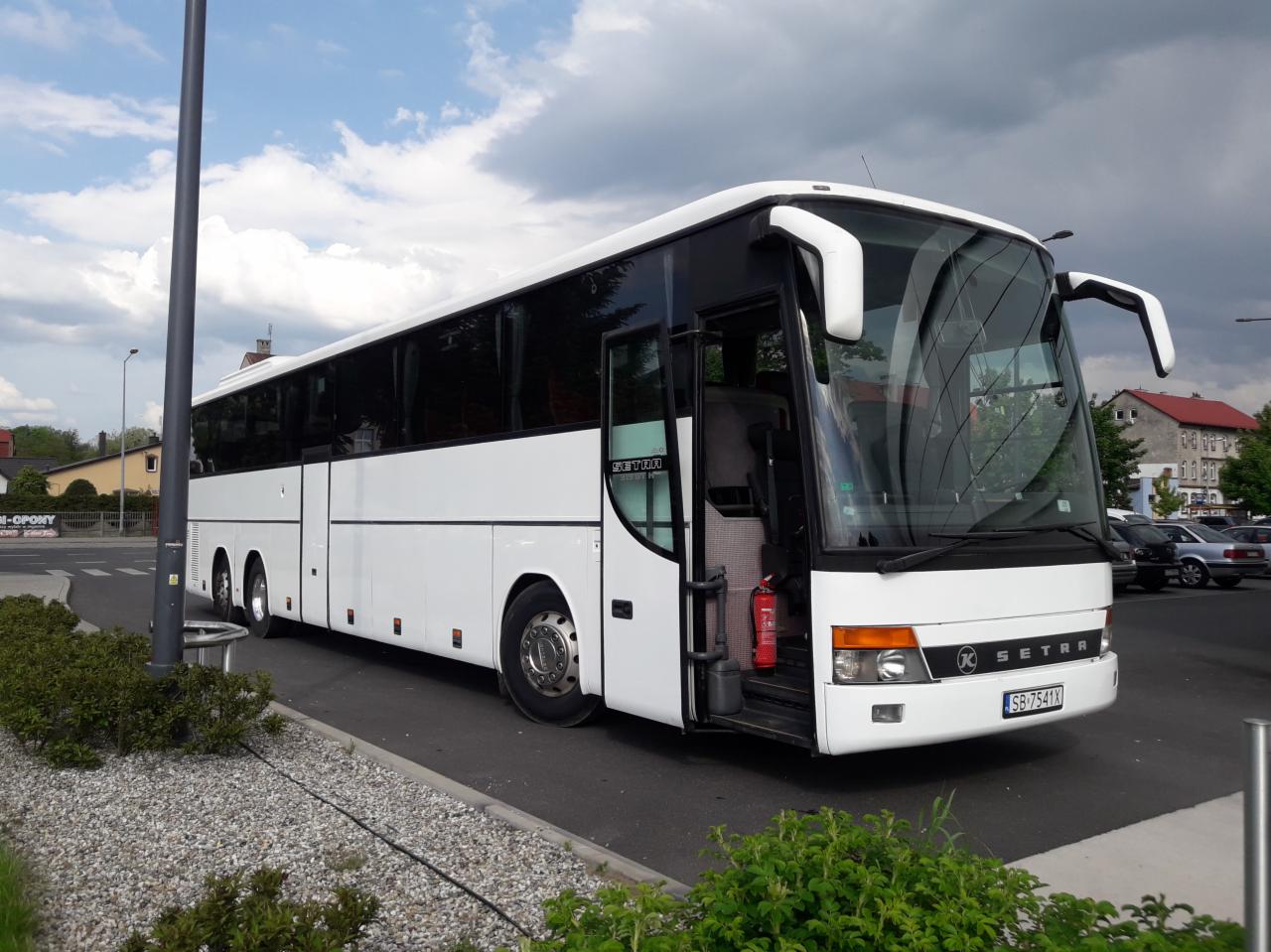 Biały autokar Setra z otwartymi drzwiami, widoczny gaśnica wewnątrz, na pierwszym planie krzewy, w tle budynki i zaparkowane samochody.