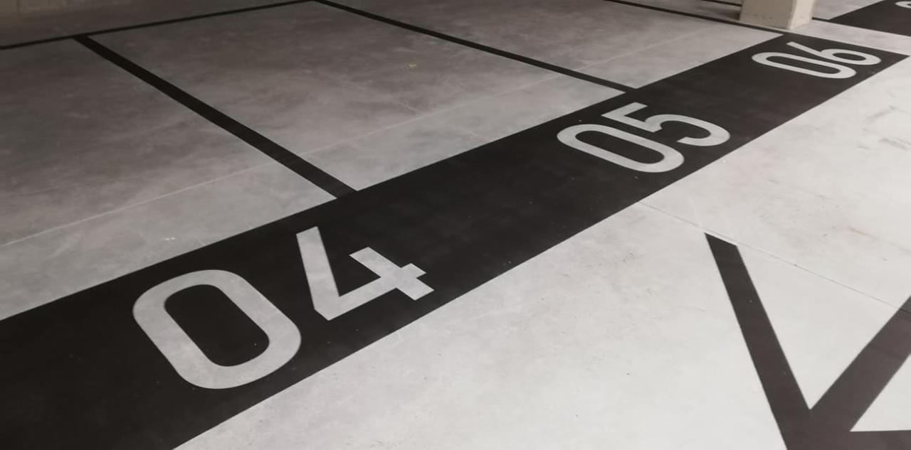 Oznakowanie miejsc parkingowych w garażu podziemnym w Krakowie: czarne pasy z białymi numerami 04, 05, 06 na szarym betonowym podłożu. Widoczne linie oddzielające miejsca postojowe.