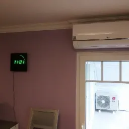 Biała jednostka klimatyzacyjna LG zamontowana nad drzwiami balkonowymi, widoczny r&oacute;wnież zegar elektroniczny na fioletowej ścianie i zewnętrzna jednostka klimatyzacyjna na balkonie.