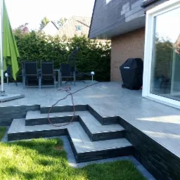 Nowoczesny taras z szarych desek kompozytowych z geometrycznymi schodami wykończonymi czarnym kamieniem, zielony trawnik, parasol i meble ogrodowe w tle.