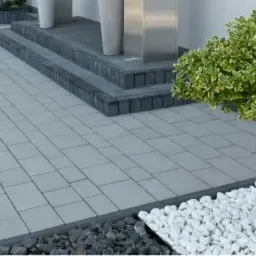 Szara kostka brukowa ułożona w regularny wzór, obrzeża z ciemniejszej kostki, dwustopniowe schody z kostki, małe drzewko w białych kamieniach, minimalistyczny design.