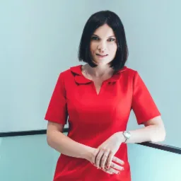 Portret kobiety w czerwonej sukience z kołnierzykiem, opierającej się o szklaną balustradę, z zegarkiem na ręku i pierścionkiem na palcu.