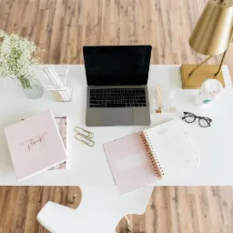 Widok z góry na białe biurko z laptopem, otwartym planerem z napisem 'hello', okularami, złotą lampką, kwiatami gipsówki w wazonie i pudełkiem 'Glamorous Planning'.