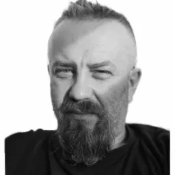 Czarno-białe portretowe zdjęcie mężczyzny z krótkim, nastroszonymi włosami i długą brodą, patrzącego wprost w obiektyw.