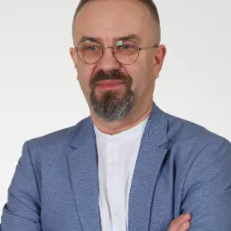 Portret mężczyzny w okularach, z brodą i wąsami, ubranego w błękitną marynarkę i białą koszulę ze stójką, z założonymi rękami na białym tle.