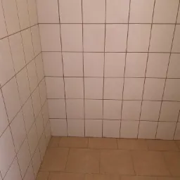 Wnętrze narożnika łazienki wyłożonego jasnymi, kwadratowymi płytkami ściennymi z ciemnymi fugami i brązowymi płytkami podłogowymi.