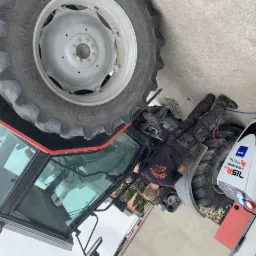 Mężczyzna w kombinezonie naprawia traktor, leżąc pod nim obok maszyny do obsługi klimatyzacji. Widoczna duża opona traktora i kabina. Ujęcie z boku, na zewnątrz.