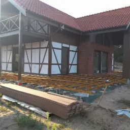 Budowa tarasu przy domu z elementami muru pruskiego i ceglaną elewacją, widoczne drewniane legary i deski tarasowe czekające na montaż.