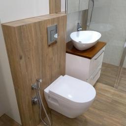 Usługi Remontowo Budowlane Krzysztof Królik - Nowoczesna łazienka z podwieszaną toaletą, bidetką i umywalką nablatową na szafce z drewnianym blatem, wykończona płytkami imitującymi drewno.