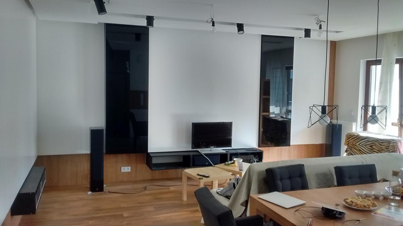 Nowoczesny salon z białymi ścianami, czarnymi panelami szklanymi, drewnianą podłogą i listwą przypodłogową, oświetleniem punktowym i wiszącymi lampami, z widocznym sprzętem RTV i meblami w tle.
