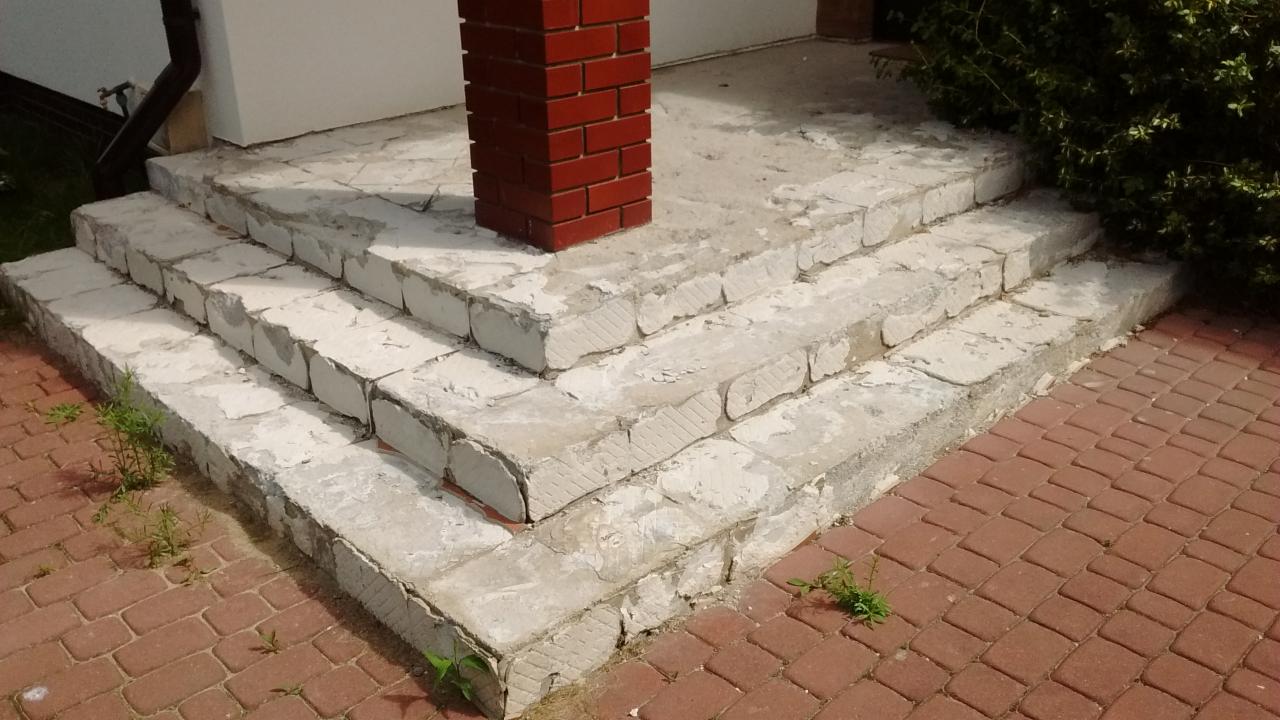 Schody wejściowe do domu, w trakcie remontu, z widocznymi śladami po starym wykończeniu, częściowo pokryte białym tynkiem, obok kolumna z czerwonej cegły i kostka brukowa.