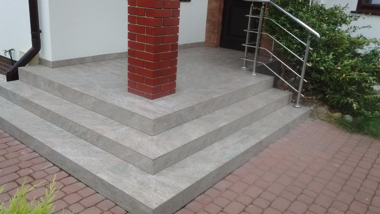 Wyłożone płytkami schody wejściowe z metalową barierką, obok kolumna z czerwonej cegły i brukowa ścieżka.