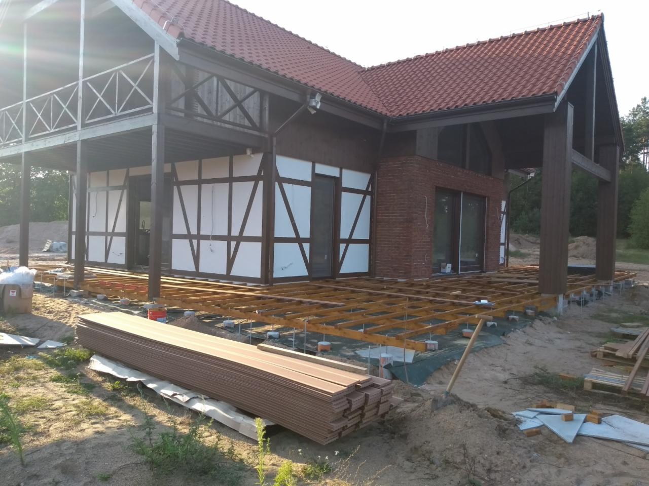 Budowa tarasu przy domu z elementami muru pruskiego i ceglaną elewacją, widoczne drewniane legary i deski tarasowe czekające na montaż.