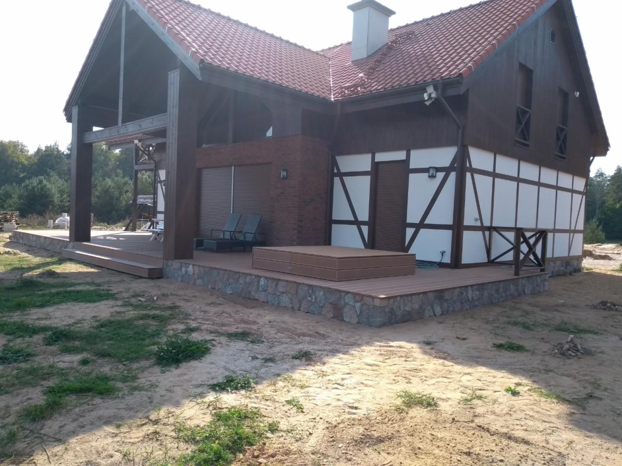 Nowoczesny dom z drewnianą elewacją szachulcową, tarasem z kompozytu i kamienną podmurówką, otoczony piaszczystym terenem.