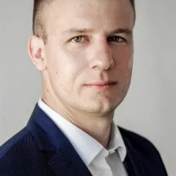 Portret mężczyzny w granatowej marynarce i białej koszuli, z krótkimi włosami i jasnymi oczami, na neutralnym tle.