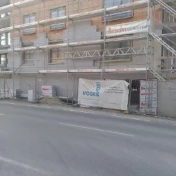 Budynek wielorodzinny w trakcie ocieplania elewacji, widoczne rusztowania, częściowo ocieplone ściany, materiały izolacyjne Flapor i baner Voges.