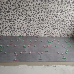 Szare płytki podłogowe w trakcie układania, z użyciem czerwonych i zielonych krzyżyków dystansowych, na tle ściany wyłożonej mozaiką z nieregularnych kamieni w odcieniach szarości i bieli.