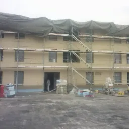 Budynek w trakcie prac elewacyjnych, osłonięty plandeką, z rusztowaniami, widoczne materiały budowlane i pracownik w kombinezonie.