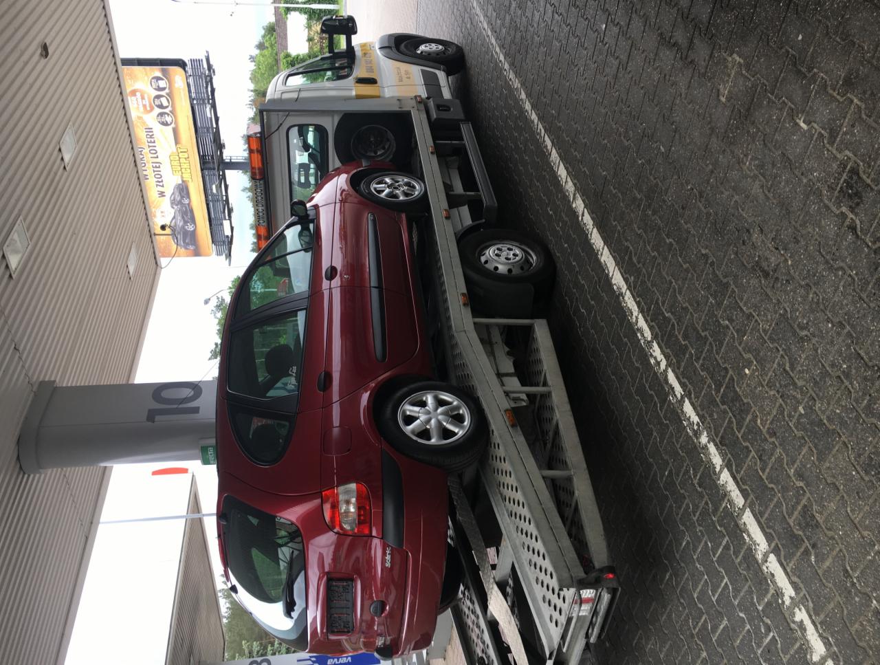 Czerwony samochód osobowy Renault Scénic załadowany na lawetę, stojący na stacji benzynowej z reklamą loterii w tle.