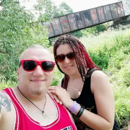 Uśmiechnięta para w okularach przeciwsłonecznych pozuje na tle zieleni i wiaduktu pokrytego graffiti. Mężczyzna w czerwonej koszulce, kobieta z dredami.