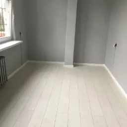 Puste, odświeżone wnętrze pokoju z szarymi ścianami i jasnymi panelami podłogowymi, widoczne gniazdko elektryczne i grzejnik żeliwny pod oknem, kolumna konstrukcyjna pomalowana na biało.