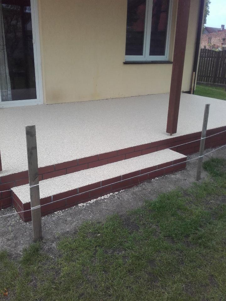 Wykończony taras z jasną, drobnokamienną powierzchnią, obrzeżony ceglanym stopniem, widok z zewnątrz.