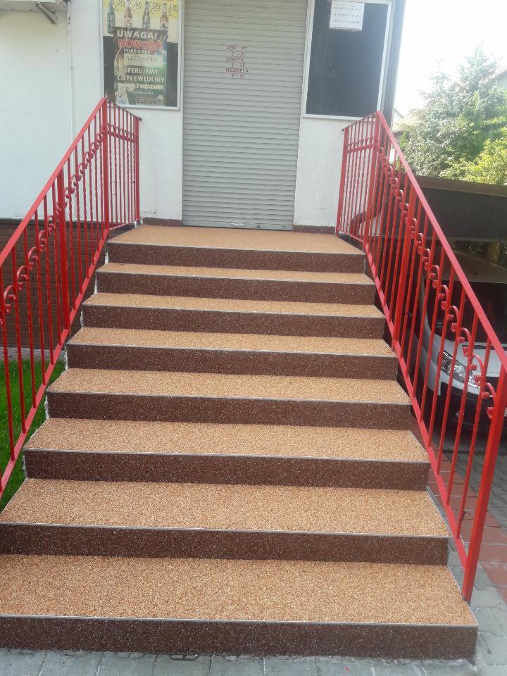 Schody wejściowe wykończone brązowo-beżową, drobną posadzką kamienną, z czerwonymi metalowymi barierkami po bokach, prowadzące do budynku z zamkniętą roletą.