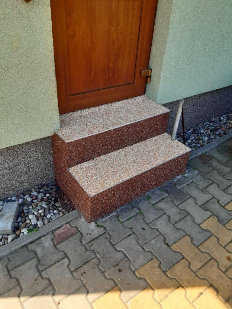 Dwa stopnie wejściowe wykończone materiałem imitującym drobny kamień, prowadzące do brązowych drzwi, z kostką brukową u podstawy.