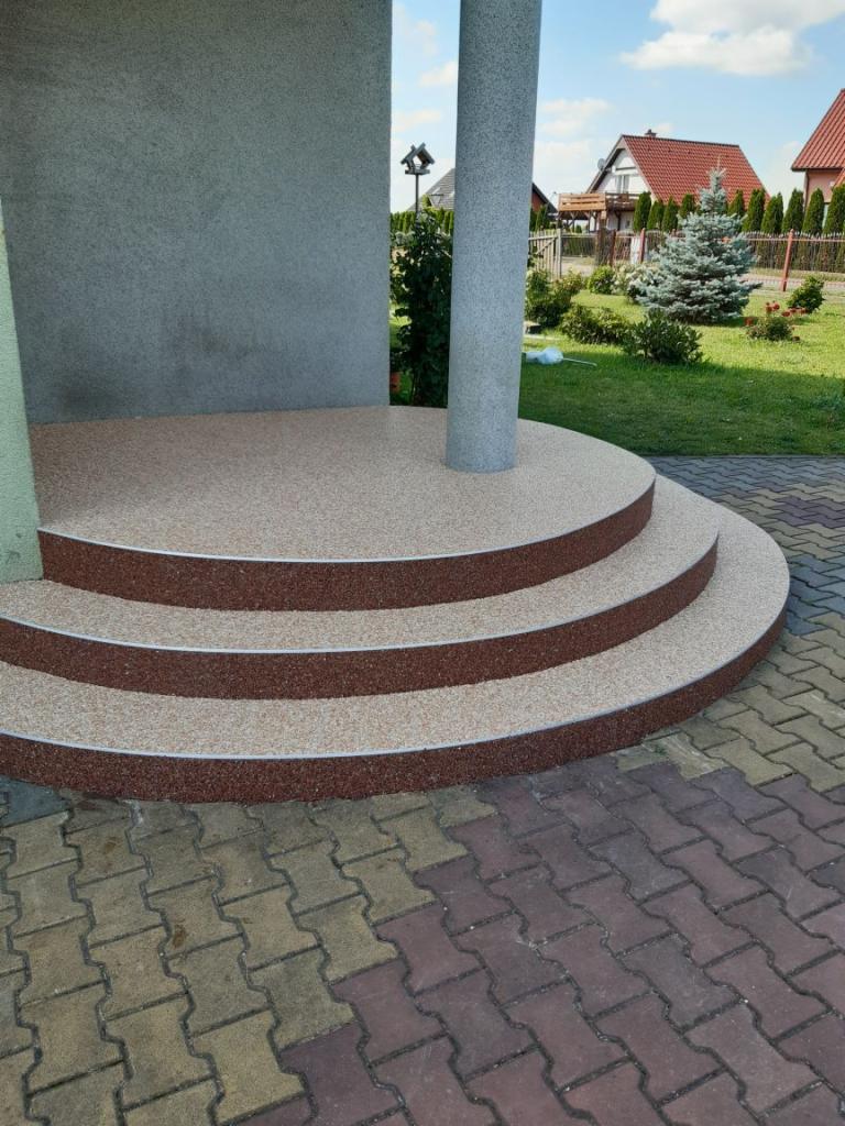 Trzy półokrągłe schody wejściowe z wykończeniem z kruszywa mineralnego w odcieniach beżu i brązu, prowadzące do kolumny podtrzymującej zadaszenie, widok z poziomu kostki brukowej.