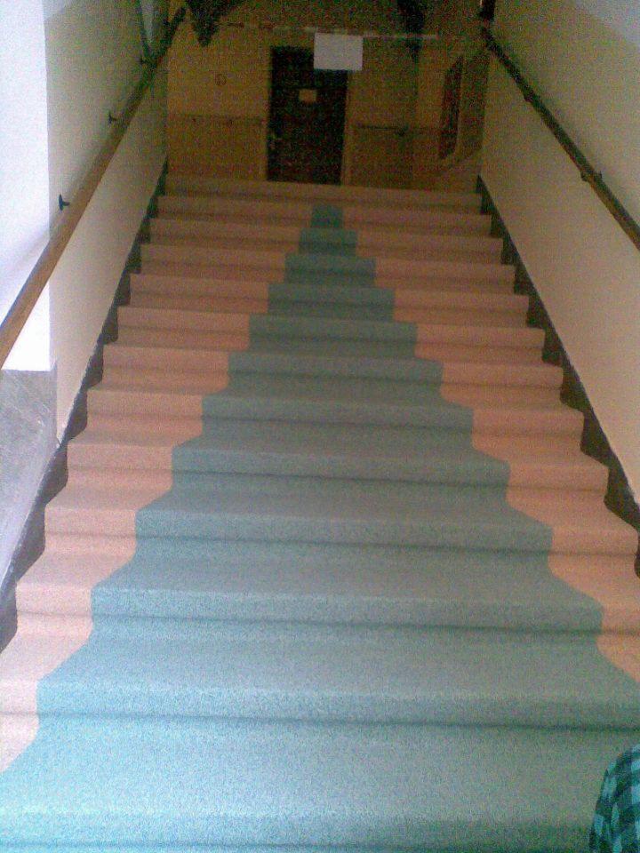 Schody pokryte szarozieloną, ziarnistą powłoką, z widoczną częścią oryginalnej, różowej powierzchni stopni po bokach, prowadzące do ciemnych drzwi.