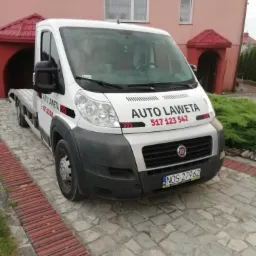 Biała laweta marki Fiat z napisem 'AUTO LAWETA' i numerem telefonu zaparkowana na kamiennym podjeździe przed budynkiem z czerwonym dachem.