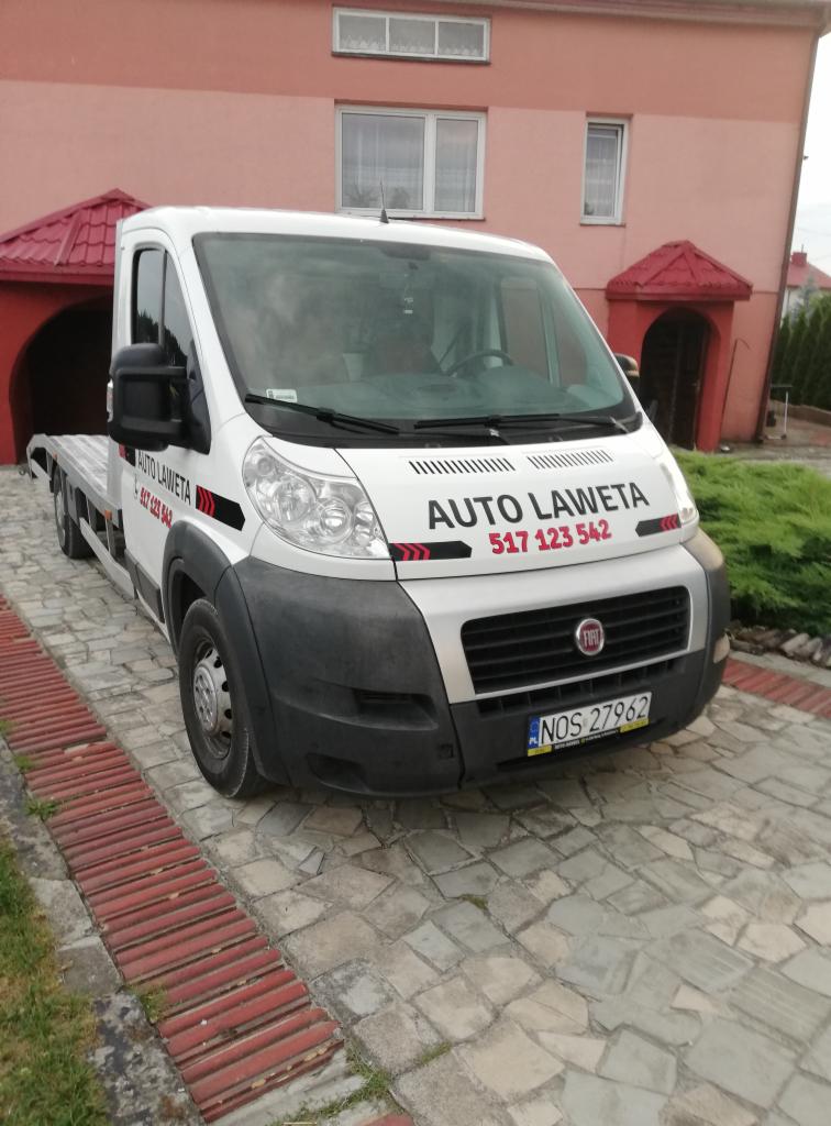 Biała laweta marki Fiat z napisem 'AUTO LAWETA' i numerem telefonu zaparkowana na kamiennym podjeździe przed budynkiem z czerwonym dachem.