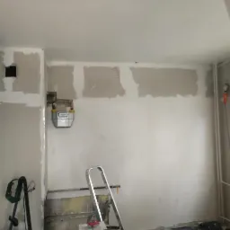 Wnętrze pokoju w trakcie remontu: ściany z widocznymi łatami gipsowymi, metalowa drabina, licznik gazowy, odkurzacz i niedokończone prace malarskie.