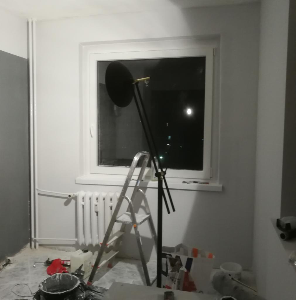 Pokój po malowaniu: świeżo pomalowane ściany na biało, drabina oparta o okno, widoczne narzędzia malarskie i wiadro z farbą na podłodze, grzejnik pomalowany na biało.