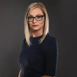 Portret kobiety w okularach, z blond włosami do ramion, ubrana w granatową sukienkę, na ciemnoszarym tle.