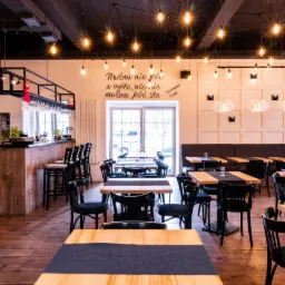 Wnętrze restauracji z drewnianymi stołami nakrytymi ciemnymi bieżnikami, czarnymi krzesłami i barem w tle, oświetlone szeregiem żar&oacute;wek Edison wiszących z sufitu, z dekoracyjnym napisem na ścianie.