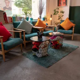 Aranżacja wnętrza z turkusowymi fotelami z drewnianymi podłokietnikami, kolorowymi poduszkami, stolikami z koszami wypełnionymi zabawkami i pluszakami, dywanikiem oraz roślinami doniczkowymi przy...