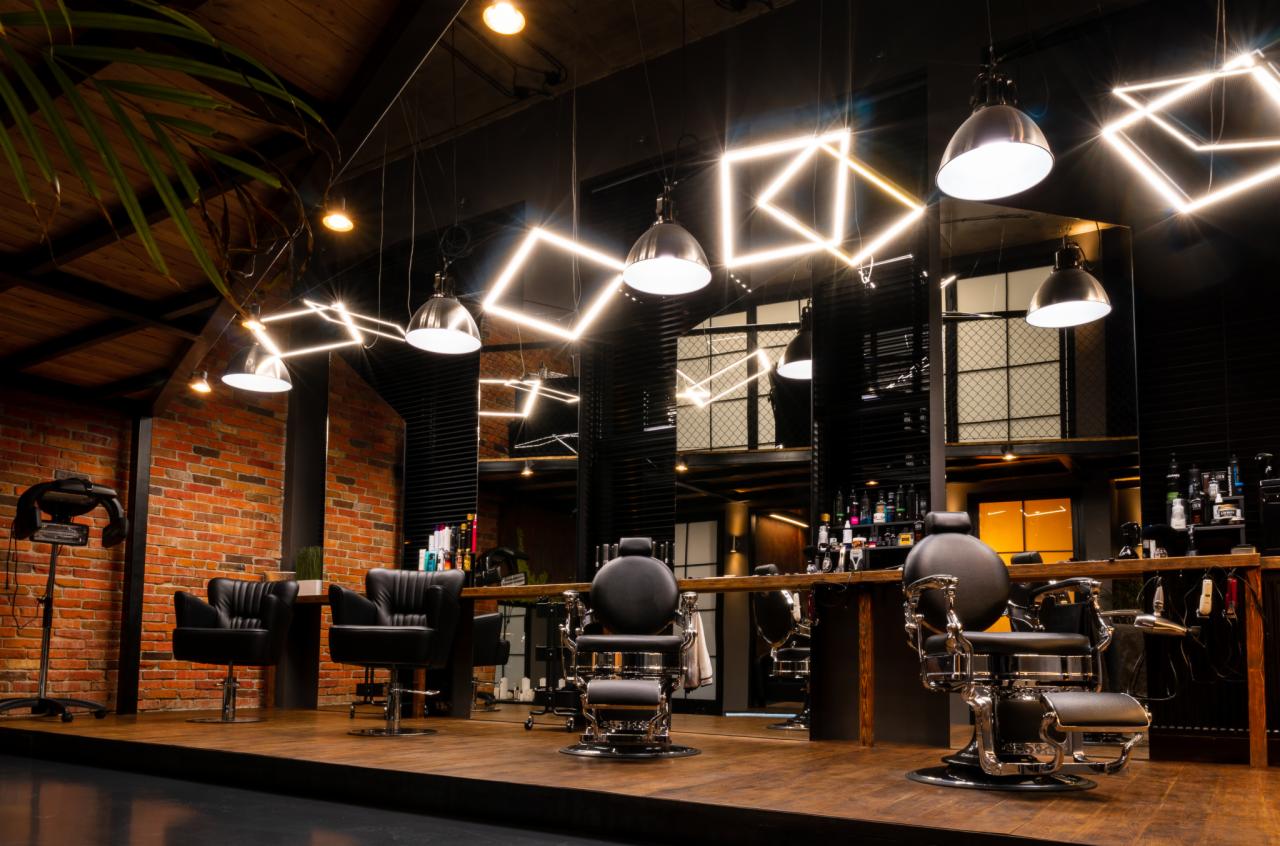 Wnętrze nowoczesnego salonu fryzjerskiego z ceglaną ścianą, stylizowanymi fotelami, lustrami i geometrycznymi lampami sufitowymi.
