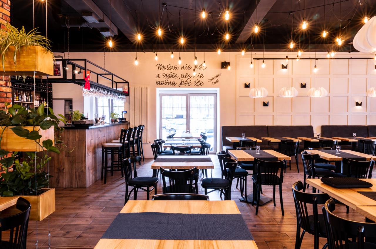 Wnętrze restauracji z drewnianymi stołami nakrytymi ciemnymi bieżnikami, czarnymi krzesłami i barem w tle, oświetlone szeregiem żarówek Edison wiszących z sufitu, z dekoracyjnym napisem na ścianie.