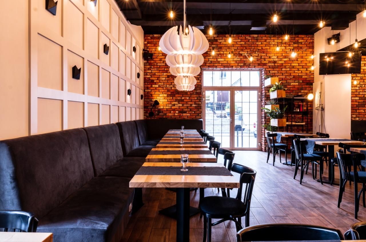 Wnętrze restauracji z długim, ciemnym narożnym siedziskiem, drewnianymi stołami z czarnymi bieżnikami i czarnymi krzesłami, ceglaną ścianą z oknem i wiszącą, wielopoziomową lampą w kształcie kwiatu.