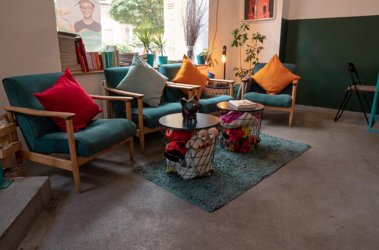 Aranżacja wnętrza z turkusowymi fotelami z drewnianymi podłokietnikami, kolorowymi poduszkami, stolikami z koszami wypełnionymi zabawkami i pluszakami, dywanikiem oraz roślinami doniczkowymi przy...