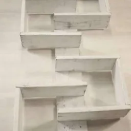 Białe, drewniane półki w kształcie schodów, przymocowane do ściany, z widocznymi śladami farby i naturalnego drewna, minimalistyczny design.