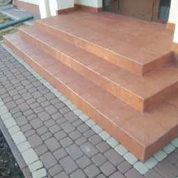 Zewnętrzne schody wejściowe wyłożone terakotą z antypoślizgowymi rowkami, obok kostka brukowa w odcieniach szarości i beżu.