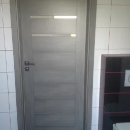 Szare drzwi wewnętrzne z poziomymi przeszkleniami i srebrną klamką w łazience wyłożonej białymi płytkami z czerwoną fugą.