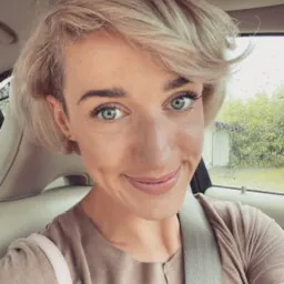 Portret uśmiechniętej kobiety z krótkimi blond włosami i zielonymi oczami, siedzącej w samochodzie w Białej Podlaskiej. Widoczny pas bezpieczeństwa i fragment krajobrazu za oknem.