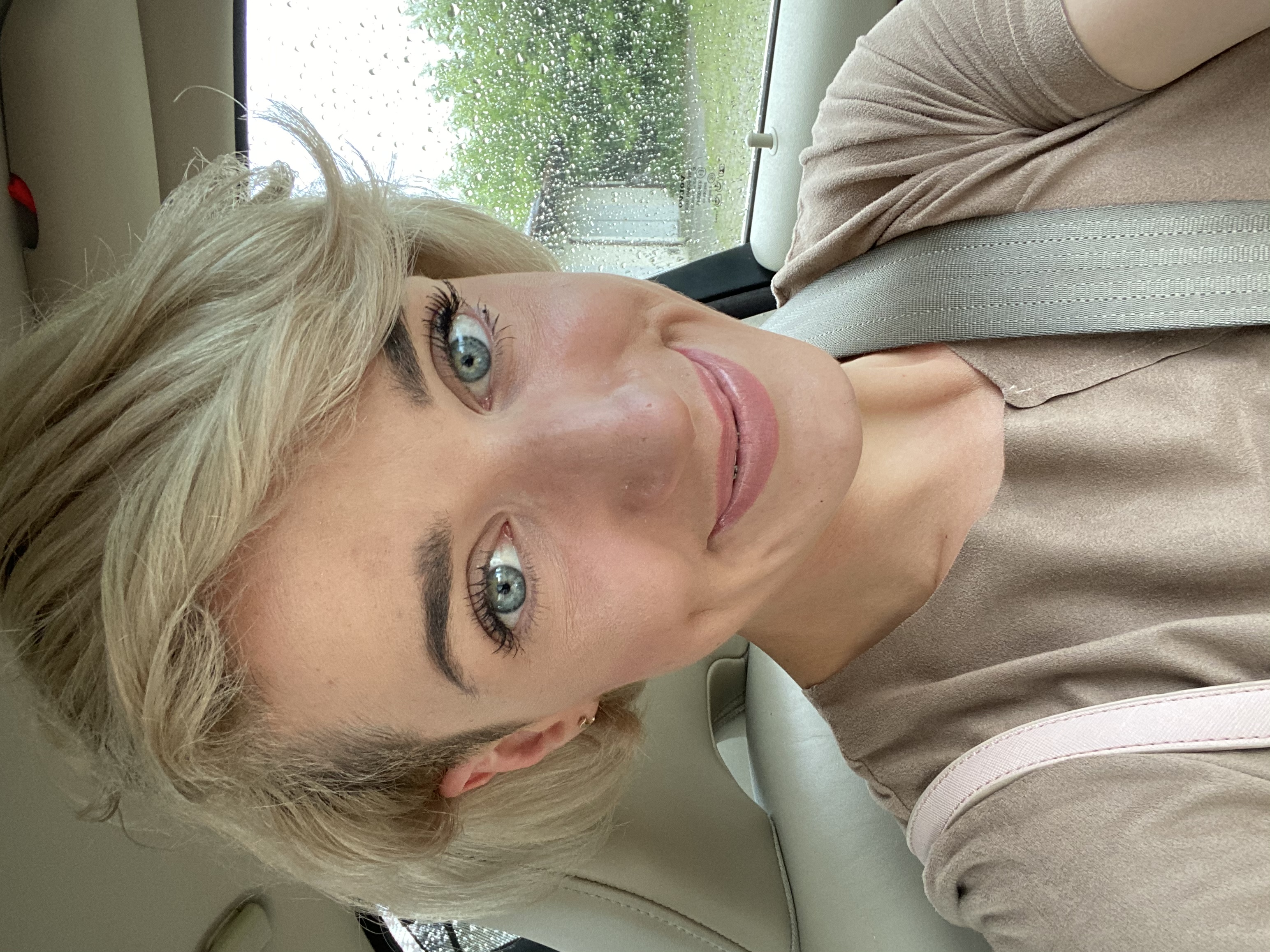 Portret uśmiechniętej blondynki w samochodzie, z widocznymi kroplami deszczu na szybie, delikatny makijaż podkreśla zielone oczy.