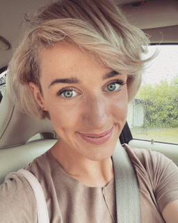 Portret uśmiechniętej kobiety z krótkimi blond włosami i zielonymi oczami, siedzącej w samochodzie w Białej Podlaskiej. Widoczny pas bezpieczeństwa i fragment krajobrazu za oknem.